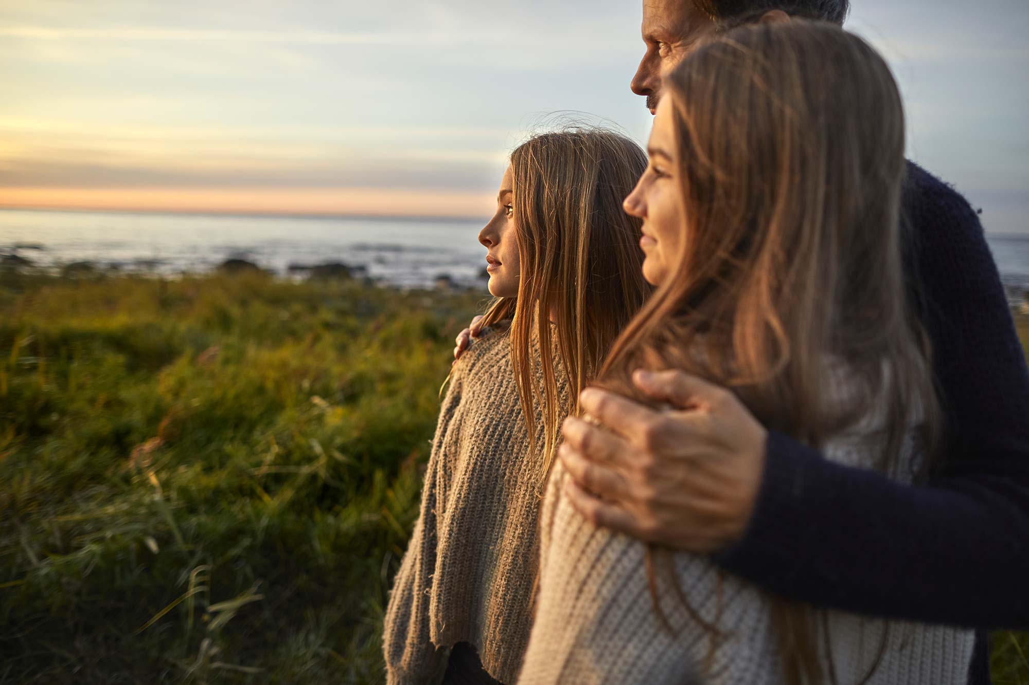 Tre personer står nära varandra och tittar ut mot ett hav där solen går ner.